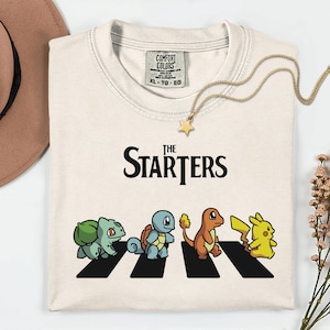 May include: Cream-coloured t-shirt with the text "The Starters" above a graphic of four cartoon characters crossing a zebra crossing. The characters are Bulbasaur, Squirtle, Charmander, and Pikachu. A gold necklace with a star pendant.