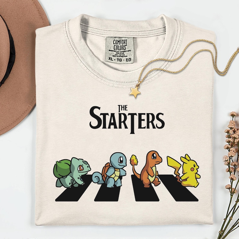 May include: Cream-coloured t-shirt with the text "The Starters" above a graphic of four cartoon characters crossing a zebra crossing. The characters are Bulbasaur, Squirtle, Charmander, and Pikachu. A gold necklace with a star pendant.