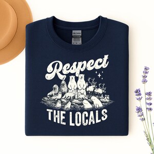 May include: A navy blue sweatshirt with the text "Respect The Locals" in a distressed white font. The graphic features arctic animals such as polar bears, walruses, and reindeer. The sweatshirt is neatly folded on a white surface.