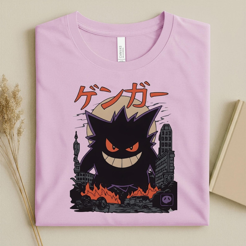 May include: A light pink t-shirt featuring a black cartoon monster with red eyes and a wide grin, standing in a city with flames and the Japanese text "ゲンガー" above. The design is centered on the shirt.