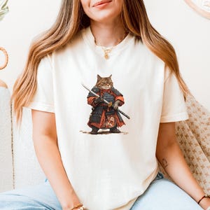May include: Cream-colored t-shirt featuring a detailed illustration of a cat dressed as a samurai warrior. The cat is wearing red and black armor and holding two swords. The design is centered on the front of the shirt.