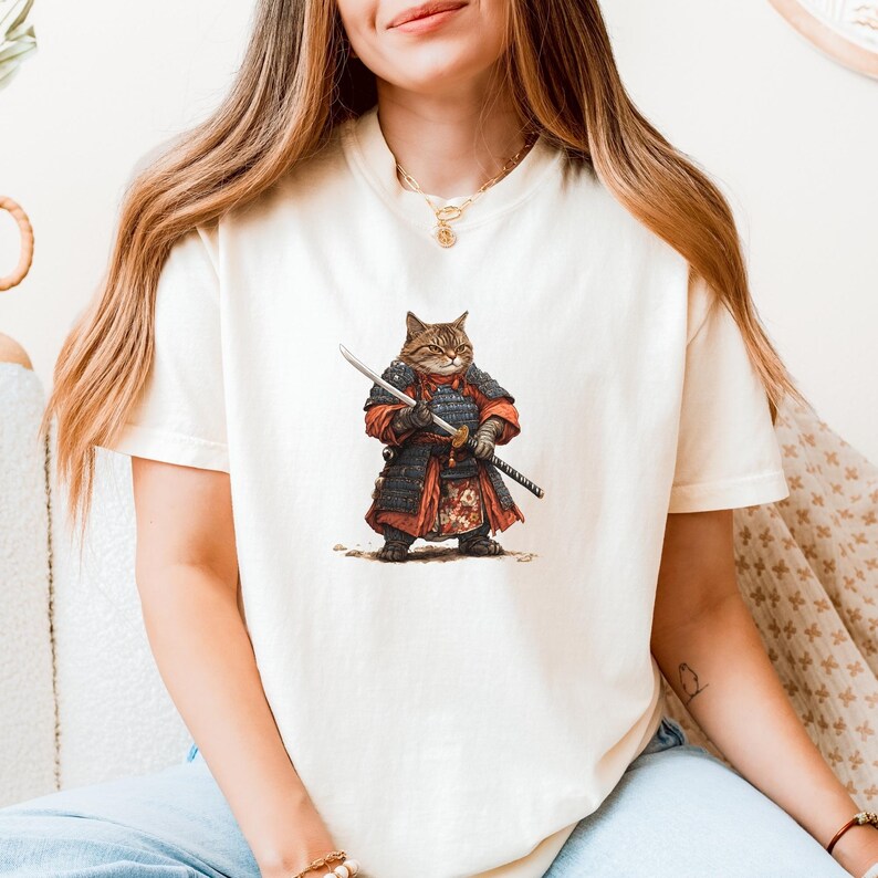 May include: Cream-colored t-shirt featuring a detailed illustration of a cat dressed as a samurai warrior. The cat is wearing red and black armor and holding two swords. The design is centered on the front of the shirt.