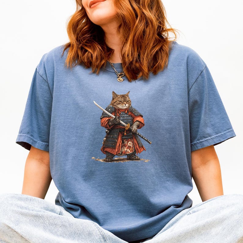May include: A blue t-shirt featuring a detailed illustration of a cat dressed as a samurai, complete with armor and a katana. The cat is brown and tan, and the armor is red and black. The shirt is a solid blue color.