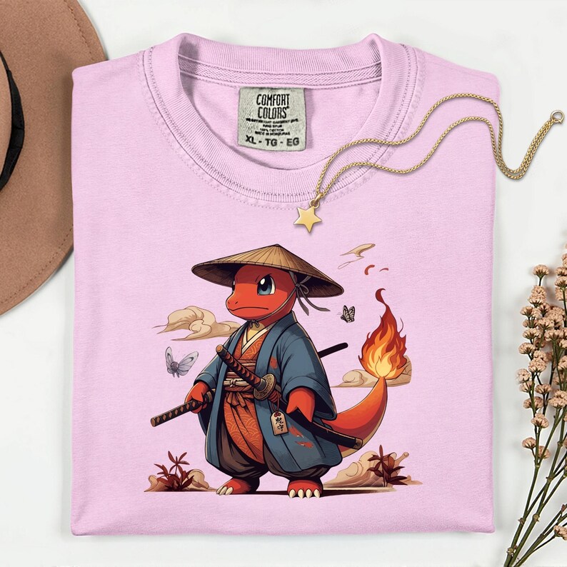 May include: A light pink Comfort Colors XL t-shirt featuring a cartoon character in a samurai outfit. The character is red with a flame tail, wearing a straw hat and holding two swords. A gold star necklace is draped across the shirt.