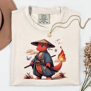 May include: Cream-colored Comfort Colors XL t-shirt featuring a cartoon red creature in a samurai outfit, holding two swords. The design includes a straw hat, blue kimono, and a flame on its tail. A gold star necklace is draped across the shirt.
