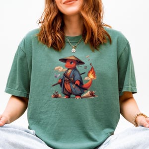 May include: Green t-shirt featuring a red cartoon character in a samurai outfit, complete with a straw hat and swords. The character has a flaming tail and is set against a backdrop of clouds and butterflies. The shirt is a solid green color.