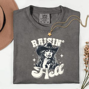 May include: Dark gray Comfort Colors t-shirt with a graphic of a woman in a cowboy hat smoking a cigarette, with the words "Raisin' Hell". A gold star pendant necklace and a sprig of pink flowers are also visible.