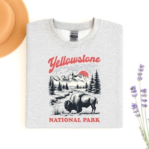 May include: A light gray sweatshirt featuring a graphic of Yellowstone National Park. The design includes a bison, mountains, trees, and the text "Yellowstone NATIONAL PARK" in red. A tan hat and lavender sprig are in the background.