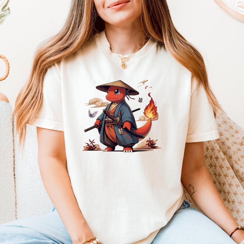 May include: Cream-colored t-shirt featuring a cartoon red creature in a samurai outfit, complete with a hat and swords. The design includes a fiery tail, butterflies, and Japanese-style clouds. A unique graphic tee.