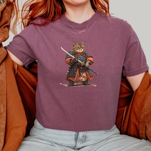 May include: A mauve-colored t-shirt featuring a detailed illustration of a cat dressed as a samurai, complete with armor and a katana. The cat is standing on a patch of dirt. The shirt has short sleeves.