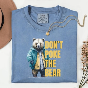 May include: A pale blue t-shirt with the yellow text "DON'T POKE THE BEAR". The shirt displays a bear wearing a jacket. A gold star pendant necklace is draped across the shirt.