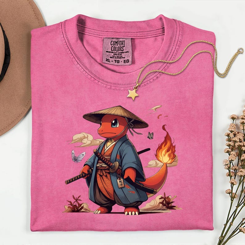 May include: A pink Comfort Colors t-shirt featuring a cartoon orange creature in a samurai outfit, complete with a straw hat and swords. The design includes butterflies and clouds. A gold star pendant necklace is draped across the shirt.