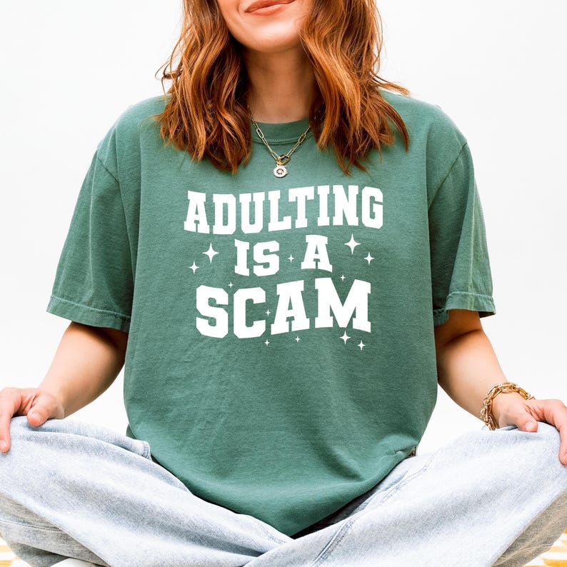 May include: A green t-shirt with the text "ADULTING IS A SCAM" in white block letters. The shirt has a relaxed fit and short sleeves. The text is accompanied by small star graphics. The person is wearing gray sweatpants.