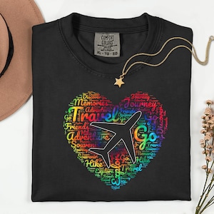 May include: Black Comfort Colors t-shirt with a heart-shaped design filled with travel-related words and a white airplane graphic. A gold necklace with a star pendant rests on the shirt. The shirt is size XL-TG-EG.