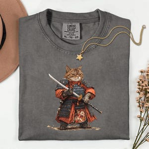 May include: A gray Comfort Colors t-shirt featuring a detailed illustration of a cat dressed as a samurai, holding a sword. The cat is wearing red and black armor. A gold star pendant necklace is draped across the shirt. The shirt size is XL-TG-EG.