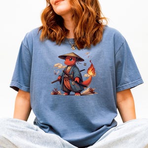 May include: Blue t-shirt featuring a cartoon character in a samurai outfit. The character is red with a flame on its tail, wearing a straw hat and holding a sword. The design includes clouds and butterflies.