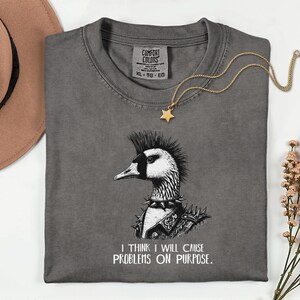 May include: A gray Comfort Colors t-shirt featuring a punk rock goose graphic with a mohawk and studded collar. The text "I THINK I WILL CAUSE PROBLEMS ON PURPOSE" is below the graphic. A gold star pendant necklace rests on the shirt.
