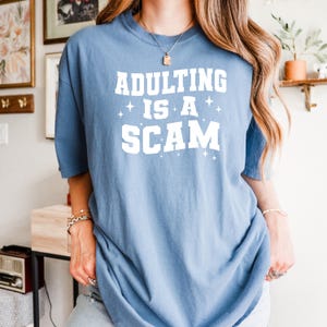 May include: A light blue t-shirt with the text "ADULTING IS A SCAM" in white block letters. The shirt has a crew neck and short sleeves. The text is centered on the front of the shirt.
