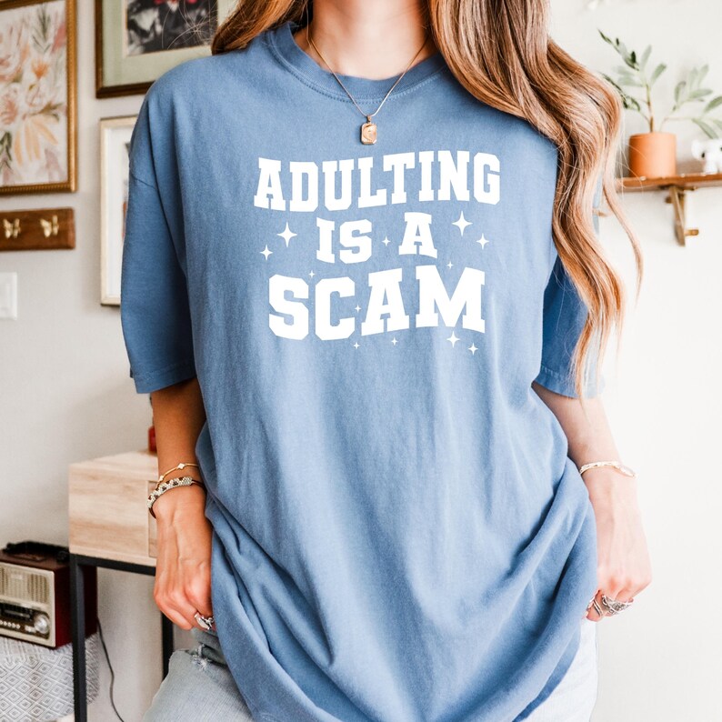 May include: A light blue t-shirt with the text "ADULTING IS A SCAM" in white block letters. The shirt has a crew neck and short sleeves. The text is centered on the front of the shirt.