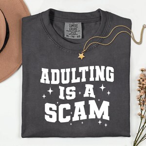 May include: Dark gray t-shirt with the phrase "ADULTING IS A SCAM" in white block letters. A gold star pendant necklace and a brown hat are also visible. The shirt is a Comfort Colors brand.