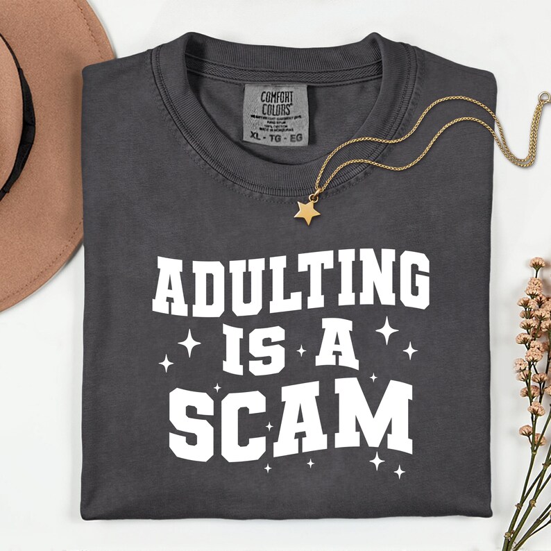 May include: Dark gray t-shirt with the phrase "ADULTING IS A SCAM" in white block letters. A gold star pendant necklace and a brown hat are also visible. The shirt is a Comfort Colors brand.