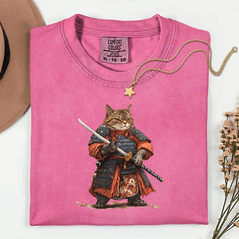 May include: A pink Comfort Colors t-shirt featuring a detailed illustration of a cat dressed as a samurai, holding a katana and a wakizashi. A gold star pendant necklace rests on the shirt. The shirt is size XL-TG-EG.