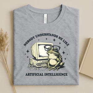 May include: A gray t-shirt featuring a graphic of a frog wearing glasses in front of a vintage computer. The text reads "NOBODY UNDERSTANDS ME LIKE ARTIFICIAL INTELLIGENCE."