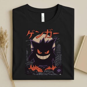 May include: Black t-shirt featuring a purple Gengar graphic with orange eyes and a wide grin, set against a cityscape with flames and the Japanese text "ゲンガー". The design includes a full moon and buildings.