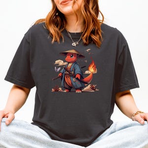 May include: Dark gray t-shirt featuring a red cartoon character in a traditional Japanese outfit, complete with a straw hat and swords. The character has a flaming tail and is set against a backdrop of clouds and butterflies. The design is detailed and colorful.