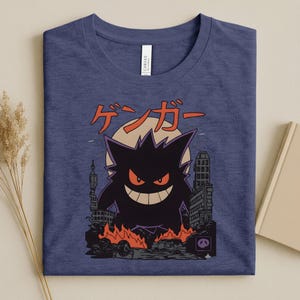 May include: A navy blue t-shirt featuring a black monster illustration with red eyes and a wide grin, set against a cityscape with flames. Japanese text is above the monster.