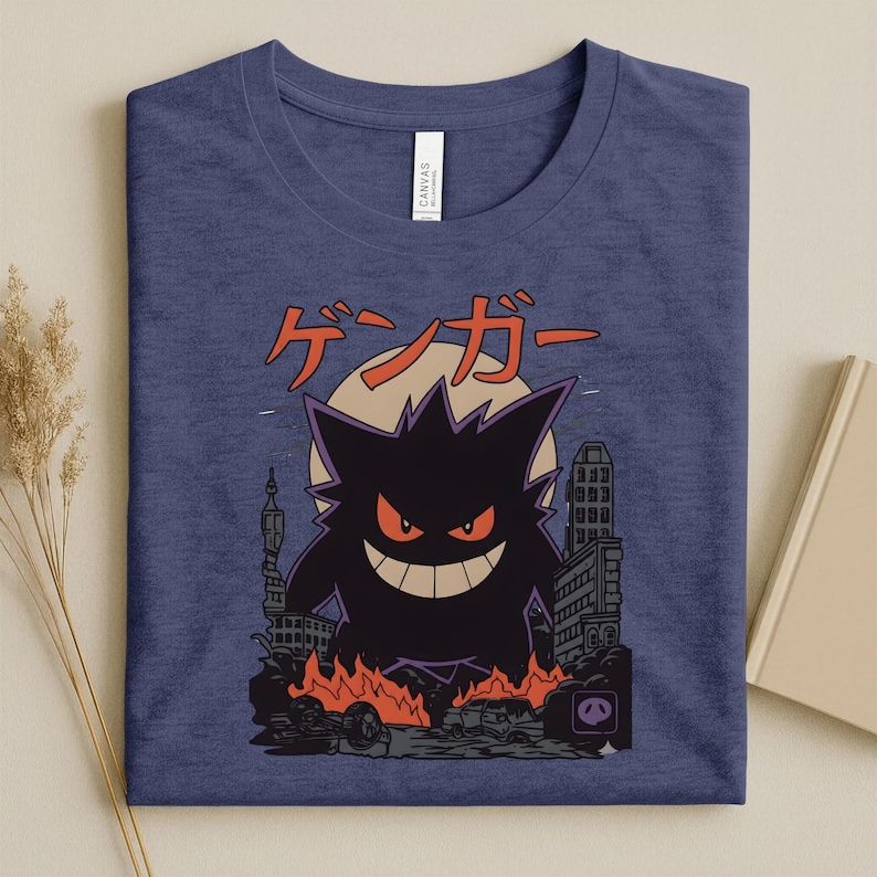May include: A navy blue t-shirt featuring a black monster illustration with red eyes and a wide grin, set against a cityscape with flames. Japanese text is above the monster.