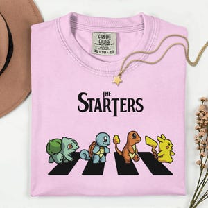 May include: Pale pink t-shirt featuring the words "The Starters" and a graphic of four cartoon characters on a zebra crossing. A gold necklace with a star pendant is on the shirt.