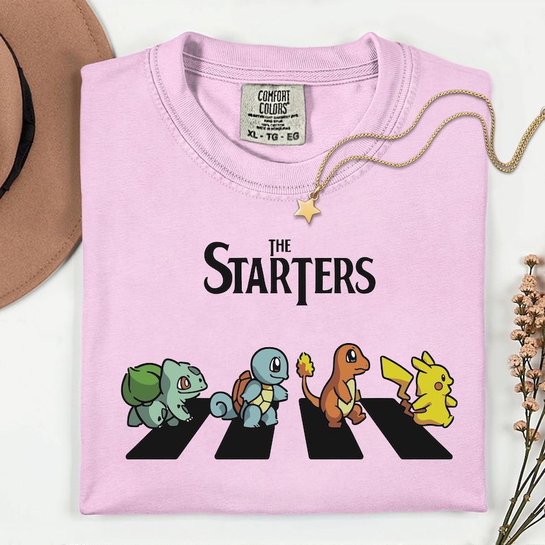 May include: Pale pink t-shirt featuring the words "The Starters" and a graphic of four cartoon characters on a zebra crossing. A gold necklace with a star pendant is on the shirt.