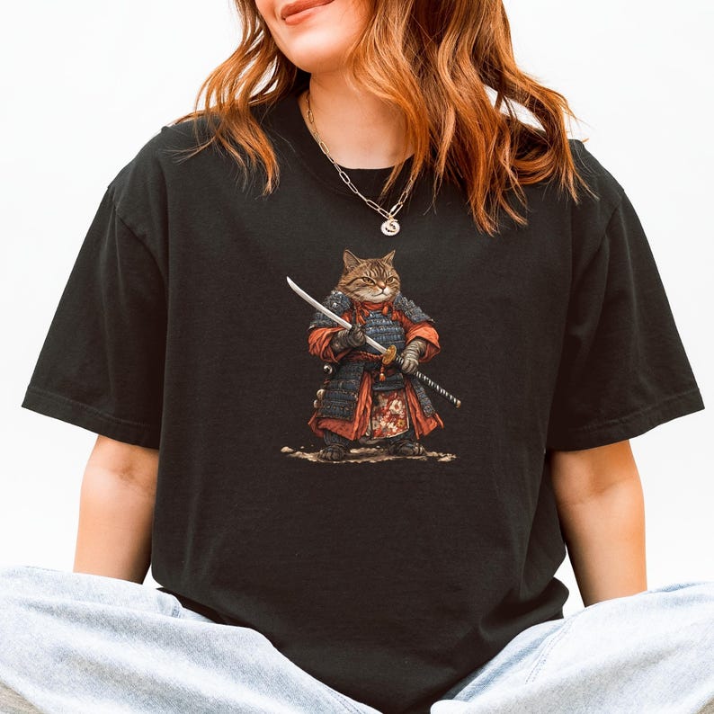 May include: Black t-shirt featuring a detailed illustration of a cat dressed as a samurai warrior, complete with armor and a katana. The cat is brown and tan, with the samurai outfit in red and black.