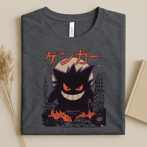 May include: Dark gray t-shirt featuring a purple and black cartoon character with red eyes and a wide grin, set against a cityscape with flames and the Japanese text "ゲンガー". The design includes a full moon.