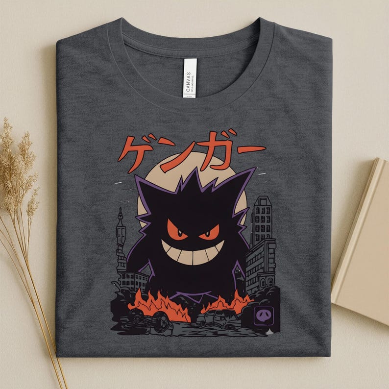 May include: Dark gray t-shirt featuring a purple and black cartoon character with red eyes and a wide grin, set against a cityscape with flames and the Japanese text "ゲンガー". The design includes a full moon.