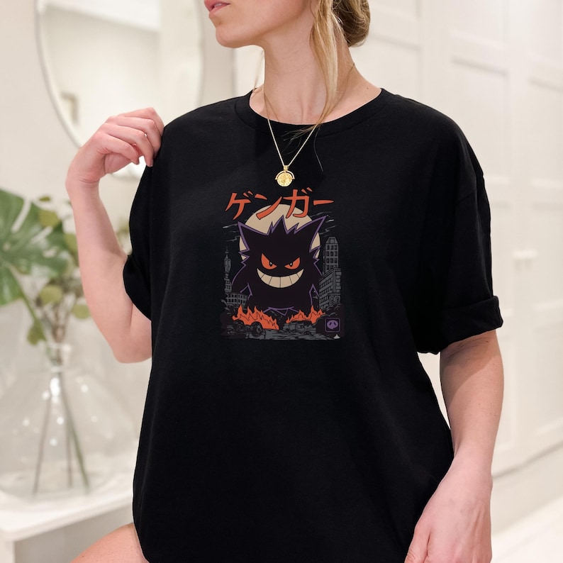 May include: Black t-shirt featuring a Gengar graphic. The design includes the Japanese characters "ゲンガー" above a silhouette of Gengar with a city backdrop and flames. The shirt is a comfortable, casual style.