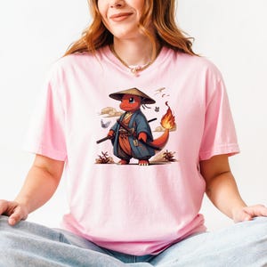 May include: A light pink t-shirt featuring a cartoon character in a samurai outfit. The character is red with a flame on its tail, wearing a blue and orange kimono, a straw hat, and carrying two swords. The design includes butterflies and clouds.