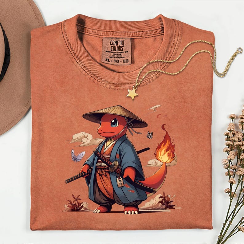 May include: Rust-colored Comfort Colors t-shirt featuring a cartoon character in a samurai outfit, complete with a straw hat and swords. The design includes clouds, butterflies, and a fiery tail. A gold star pendant necklace rests on the shirt.