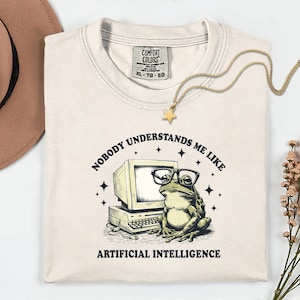 May include: Cream-colored t-shirt with a graphic of a frog wearing glasses next to a computer. Text reads "NOBODY UNDERSTANDS ME LIKE ARTIFICIAL INTELLIGENCE." A gold necklace with a star pendant is draped on the shirt.