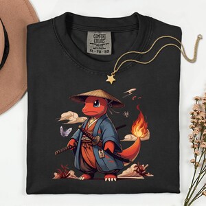 May include: Black Comfort Colors t-shirt featuring a cartoon character in a samurai outfit, holding two swords. The character is red with a flame tail and wears a straw hat and blue kimono. A gold star pendant necklace is draped across the shirt.