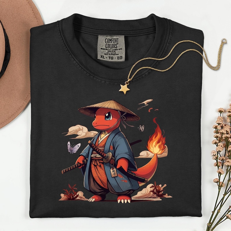 May include: Black Comfort Colors t-shirt featuring a cartoon character in a samurai outfit, holding two swords. The character is red with a flame tail and wears a straw hat and blue kimono. A gold star pendant necklace is draped across the shirt.