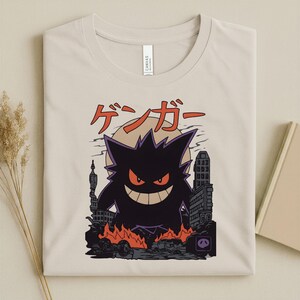 May include: Beige t-shirt featuring a black Gengar graphic with red eyes and a wide grin, set against a cityscape with flames. Japanese text is above the graphic. The shirt is folded.