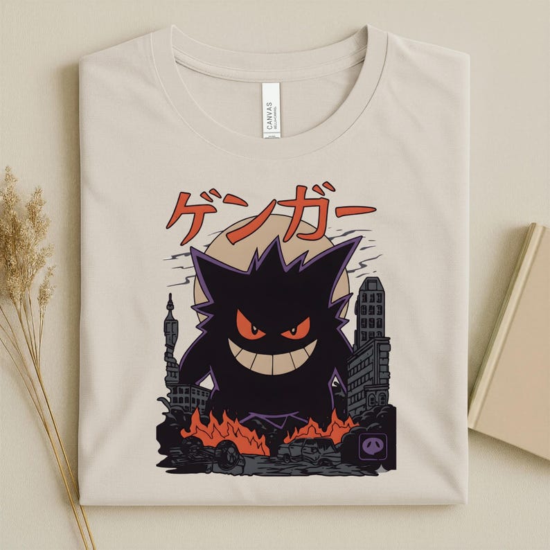 May include: Beige t-shirt featuring a black Gengar graphic with red eyes and a wide grin, set against a cityscape with flames. Japanese text is above the graphic. The shirt is folded.