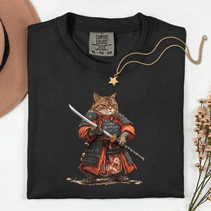 May include: Black Comfort Colors t-shirt featuring a cartoon cat dressed as a samurai, holding two swords. The cat is wearing red and black armor. A gold star pendant necklace and a tag that says "COMFORT COLORS" are also visible.