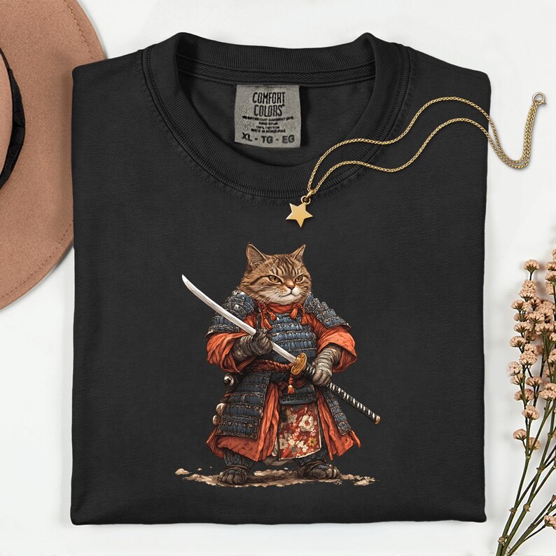 May include: Black Comfort Colors t-shirt featuring a cartoon cat dressed as a samurai, holding two swords. The cat is wearing red and black armor. A gold star pendant necklace and a tag that says "COMFORT COLORS" are also visible.