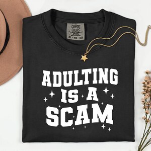 May include: Black t-shirt with the phrase "ADULTING IS A SCAM" in white block letters. A gold star pendant necklace and a brown hat are also visible. The shirt has a Comfort Colors tag.