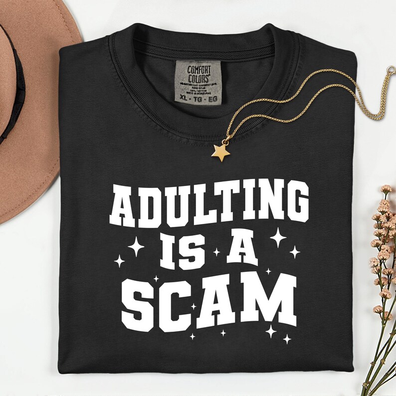 May include: Black t-shirt with the phrase "ADULTING IS A SCAM" in white block letters. A gold star pendant necklace and a brown hat are also visible. The shirt has a Comfort Colors tag.