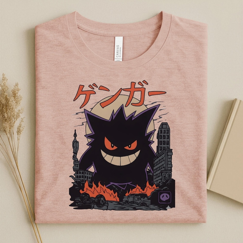 May include: Pink t-shirt featuring a black graphic of a monster with red eyes and a wide grin, destroying a city. Japanese text is above the monster. The design includes flames, buildings, and a full moon.