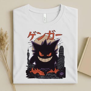 May include: White t-shirt featuring a black cartoon monster with red eyes and a wide grin, standing in a city with flames and the Japanese text "ゲンガー" above. The design is a graphic illustration.